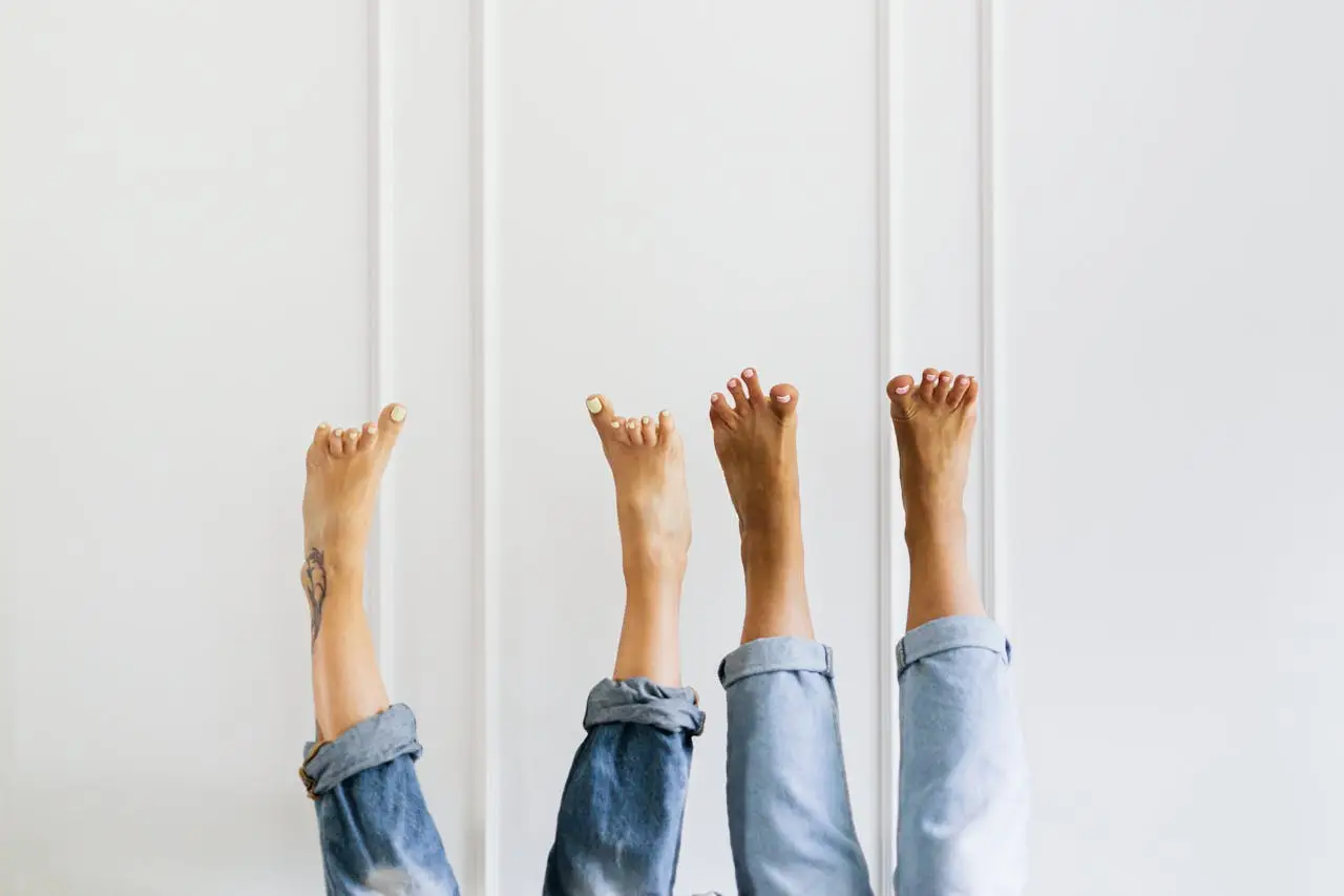 hero-img-01 Three pairs of bare feet playfully raised against a white wall, wearing casual denim jeans.